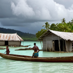 **

Prompt: A dramatic, eye-level shot of a coastal village in Vanuatu being inundated by rising sea levels. Dilapidated houses are partially submerged, with concerned villagers wading through the water, carrying belongings. A traditional outrigger canoe is visible. The sky is overcast and stormy. Emphasize the vulnerability and the immediate threat of climate change. Style: Photojournalistic, realistic.

**